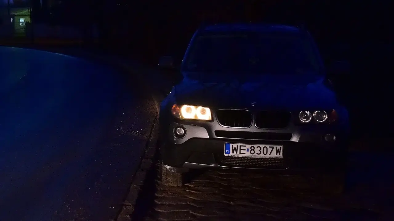 Ciemny SUV z włączonymi światłami pozycyjnymi stoi na mokrej, brukowanej drodze.