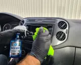 Ręce w czarnych rękawiczkach czyszczą deskę rozdzielczą samochodu. Pokazano, jak wypolerować plastik domowym sposobem, używając płynu "Wash Mist".