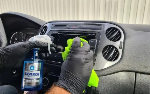 Ręce w czarnych rękawiczkach czyszczą deskę rozdzielczą samochodu, używając płynu "Wash Mist". To prosty sposób, jak pomalować plastiki w samochodzie, nadając im nowy wygląd.