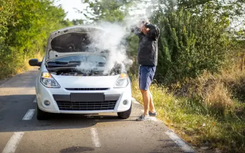 Mężczyzna z rozpaczą w rękach przy białym samochodzie, z którego bucha dym. Rozbiegany diesel na poboczu drogi.