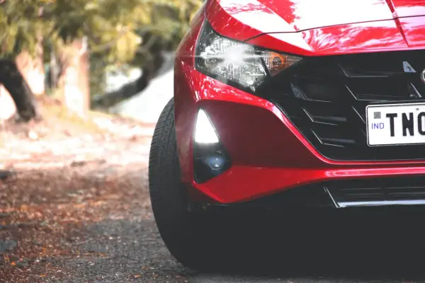 Czerwony Hyundai i20 z zapalonymi światłami dziennymi i reflektorami. Widać fragment maski, grill i tablicę rejestracyjną.