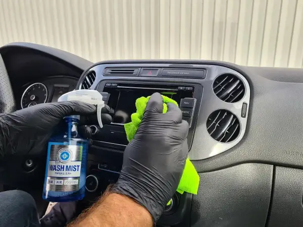 Ręce w czarnych rękawiczkach czyszczą deskę rozdzielczą samochodu. Pokazano, jak wypolerować plastik domowym sposobem, używając płynu "Wash Mist".