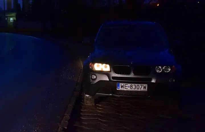 Ciemny SUV z włączonymi światłami pozycyjnymi stoi na mokrej, brukowanej drodze.