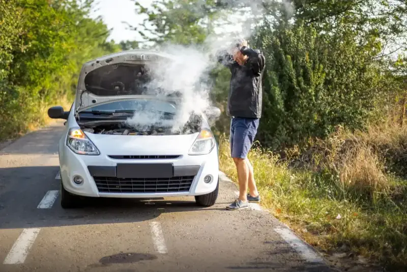Mężczyzna z rozpaczą w rękach przy białym samochodzie, z którego bucha dym. Rozbiegany diesel na poboczu drogi.